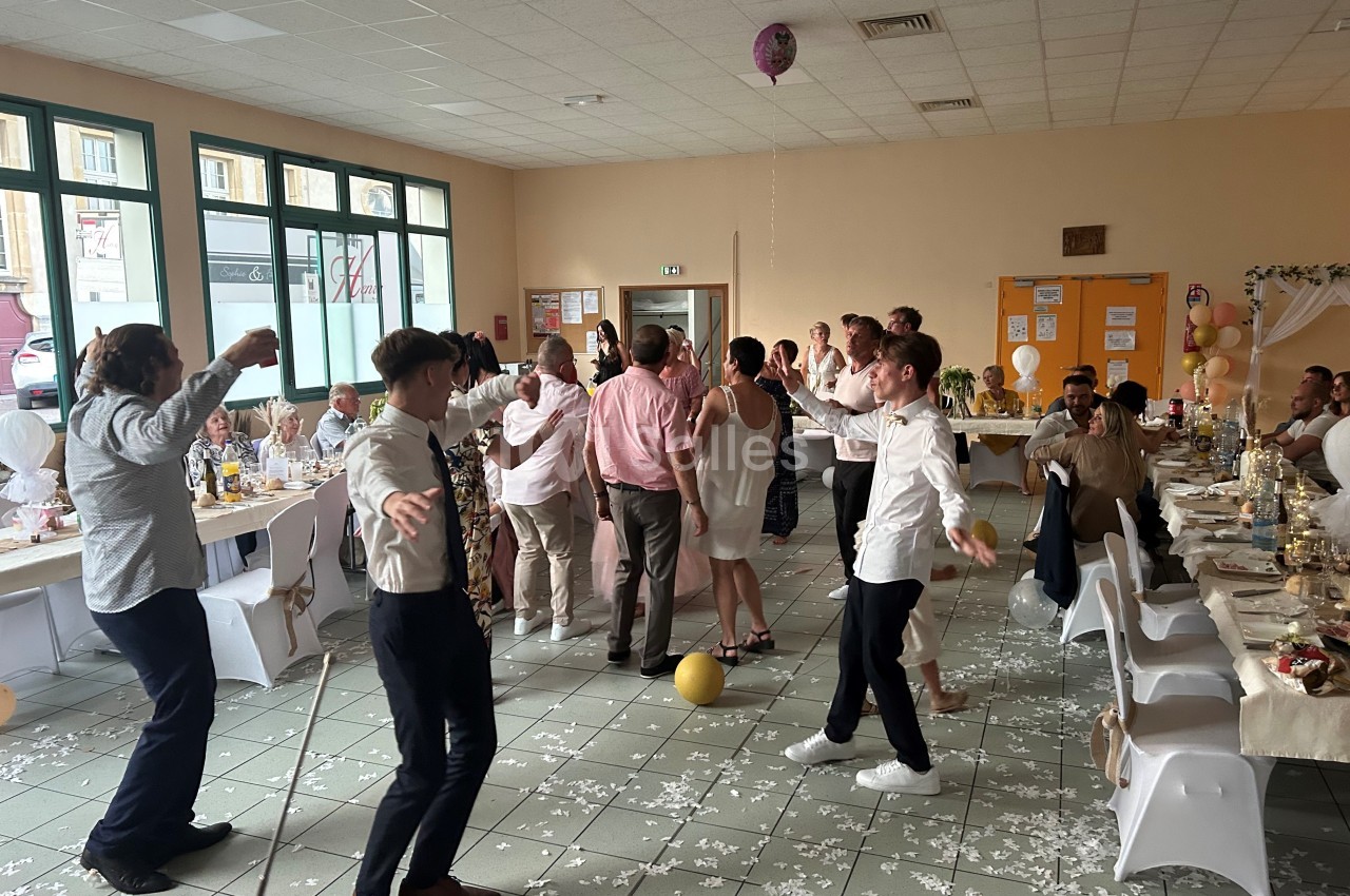 Des invités dansent dans une salle décorée pour une réception, avec des tables dressées et des ballons au sol.