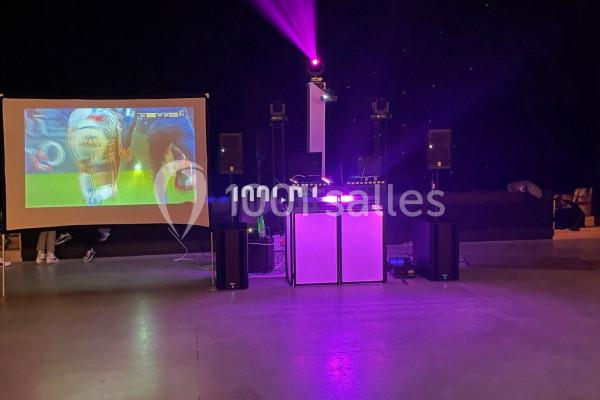 Écran projetant un match de football à côté d'une installation de DJ avec éclairage violet dans une salle sombre.