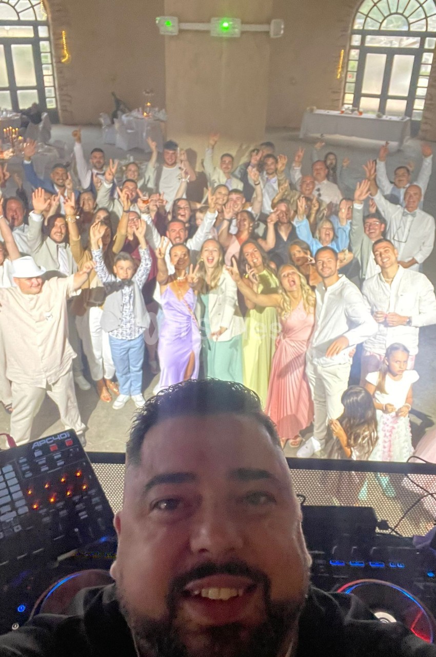 Un DJ prend un selfie devant un groupe de personnes souriantes et levant les bras dans une salle de réception lumineuse.