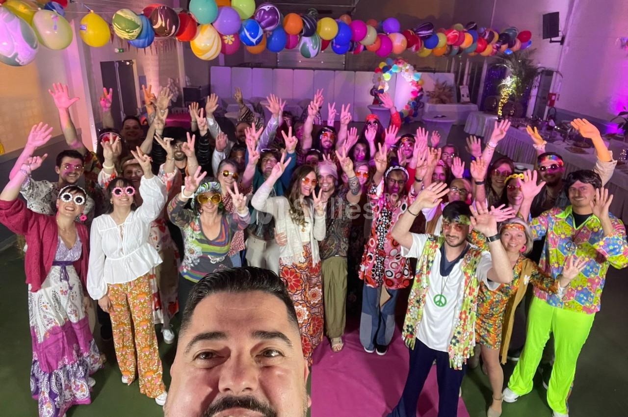 Un groupe de personnes en tenue colorée lève les bras dans une salle décorée de ballons lors d'un événement festif.