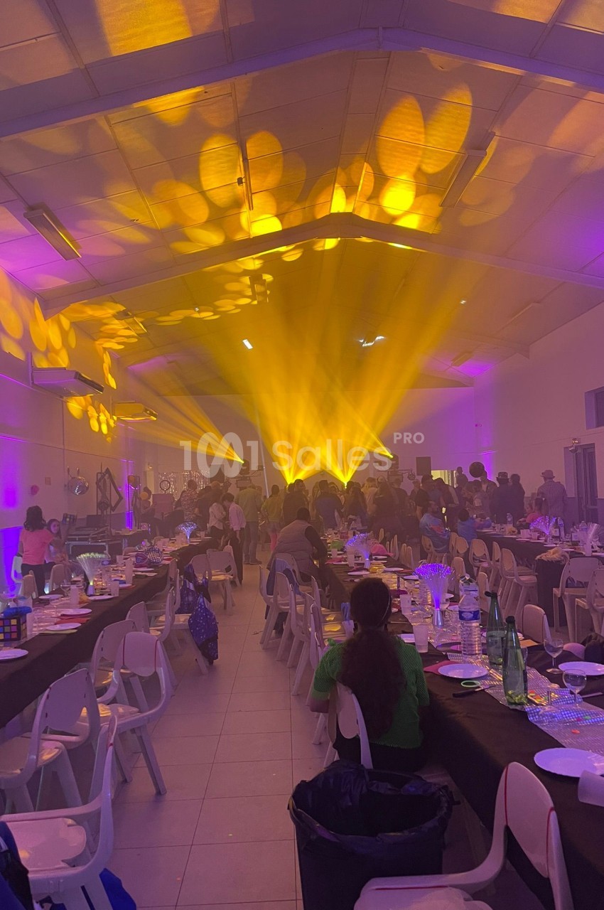 Salle de réception animée avec des invités assis, éclairage jaune vif et décorations sur les tables.