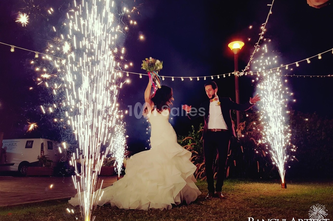 Un couple en tenue de mariage danse en extérieur, entouré de fontaines d'étincelles et de guirlandes lumineuses.