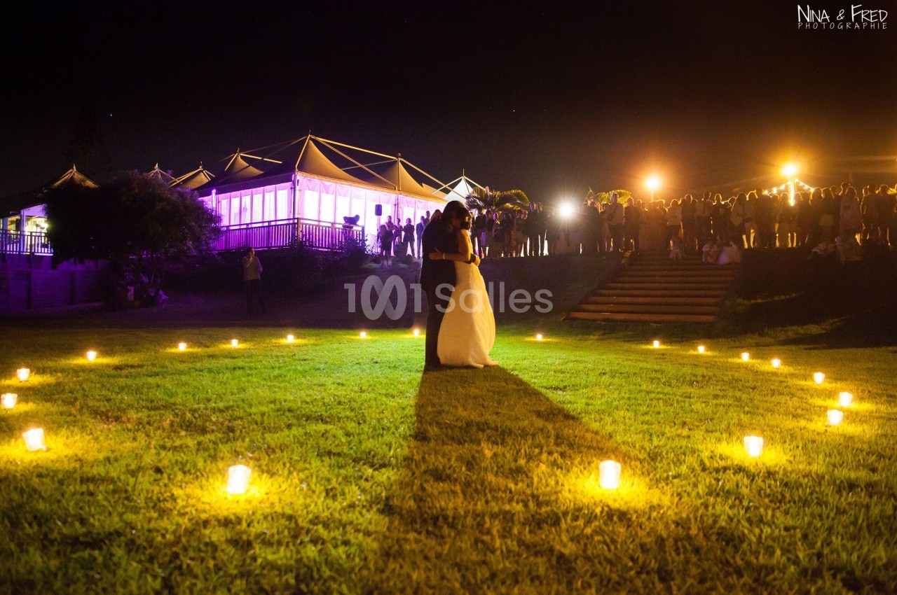 Un couple danse au centre d'une pelouse éclairée par des bougies, avec un bâtiment illuminé et des invités en arrière-plan.