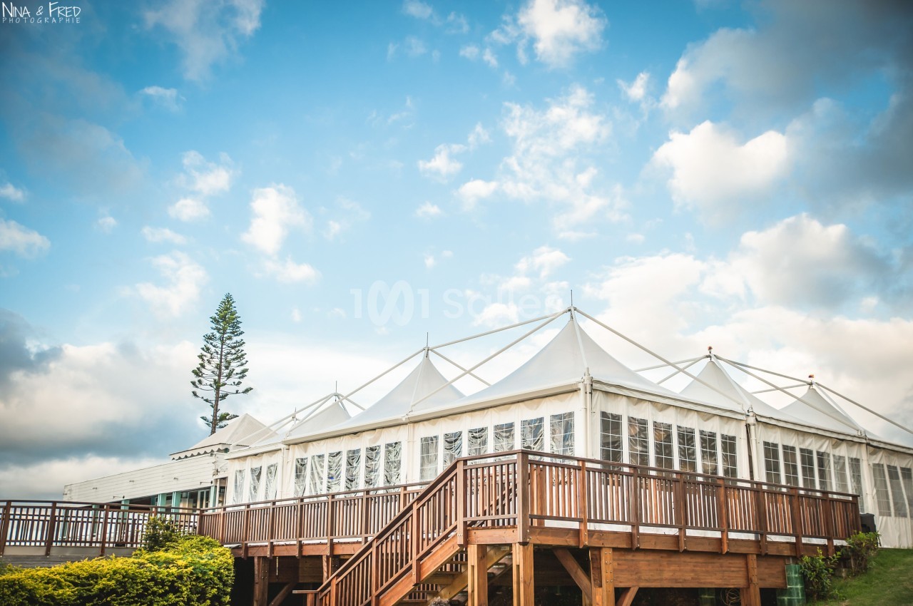 Grande structure blanche avec toit en toile, entourée de verdure et d'une terrasse en bois, sous un ciel partiellement…