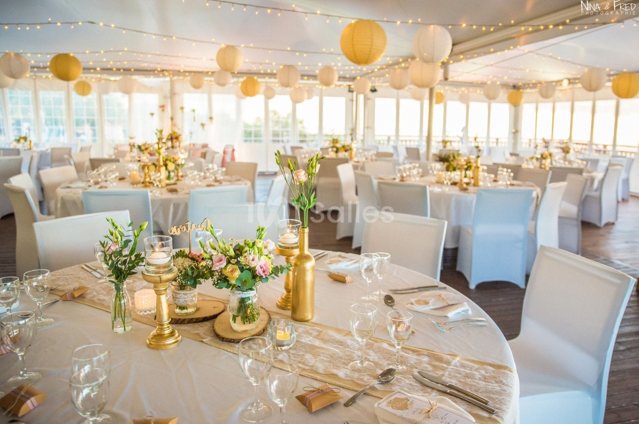 Salle de réception décorée avec des tables rondes, nappes blanches, centres de table floraux et lanternes suspendues.