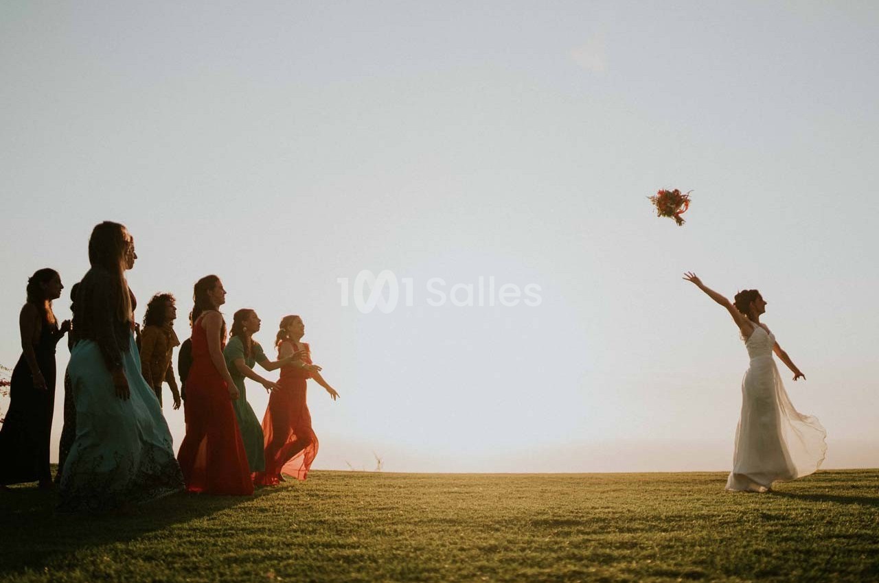 Une mariée lance son bouquet vers un groupe de femmes sur une pelouse au coucher du soleil.