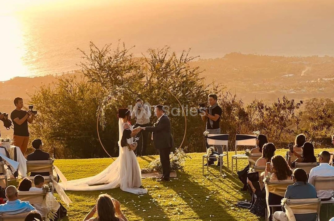 Un couple échange ses vœux de mariage en plein air au coucher du soleil, entouré d'invités assis.
