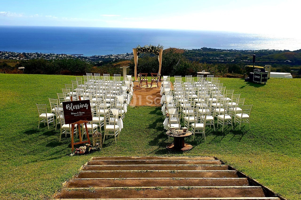 Chaises blanches disposées en rangées sur une pelouse, face à une arche décorée, avec vue sur la mer et une ville en arrière…