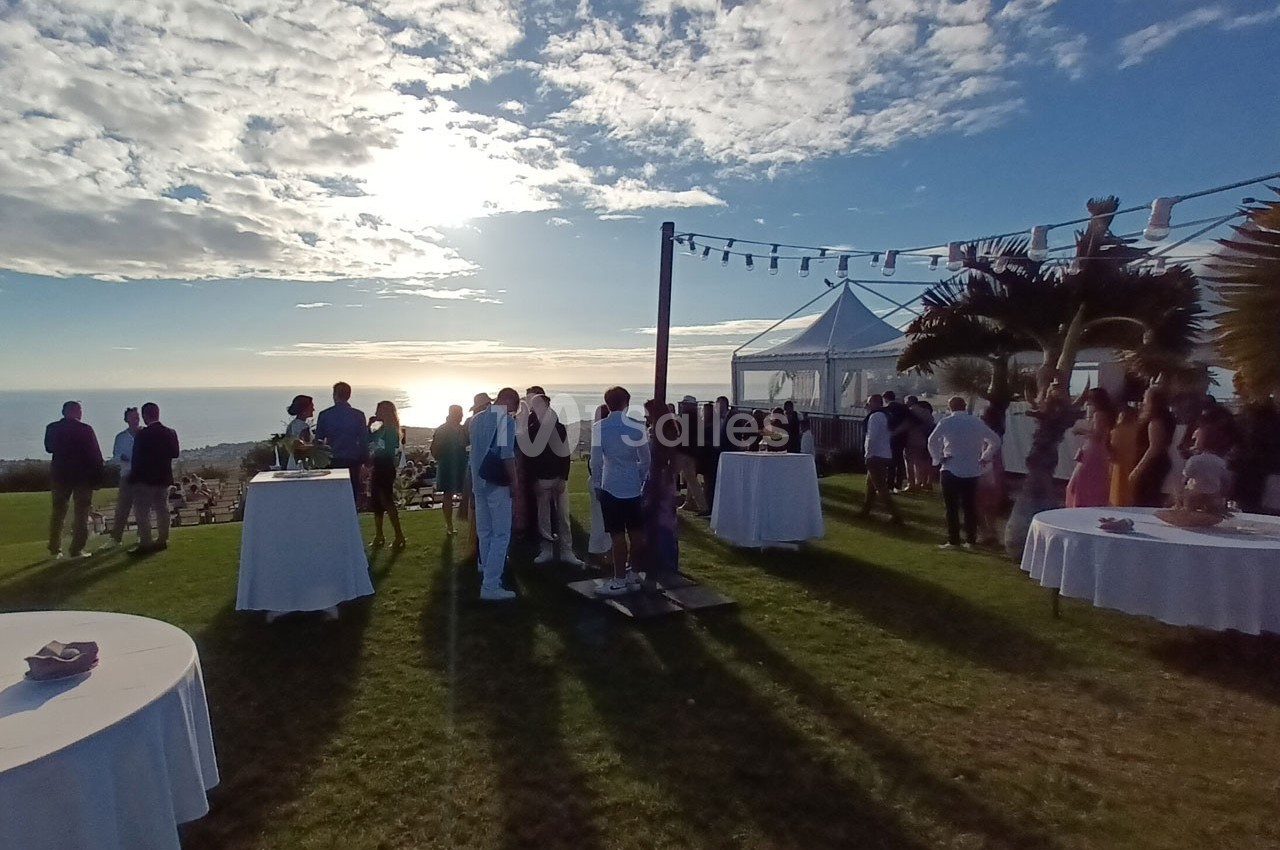 Groupe de personnes rassemblées sur une pelouse en bord de mer au coucher du soleil, avec des tables et une tente blanche.