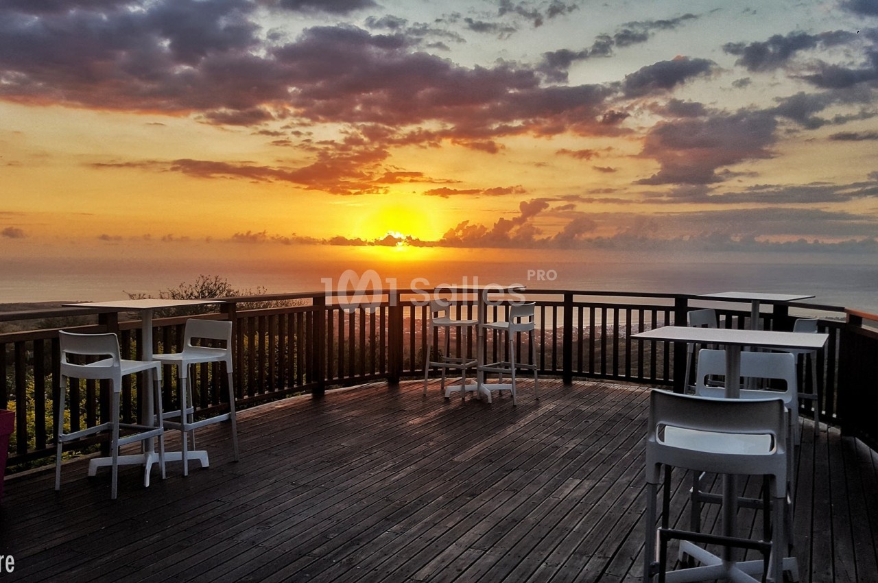Terrasse en bois avec des tables et chaises blanches, offrant une vue sur un coucher de soleil au-dessus de l'océan.