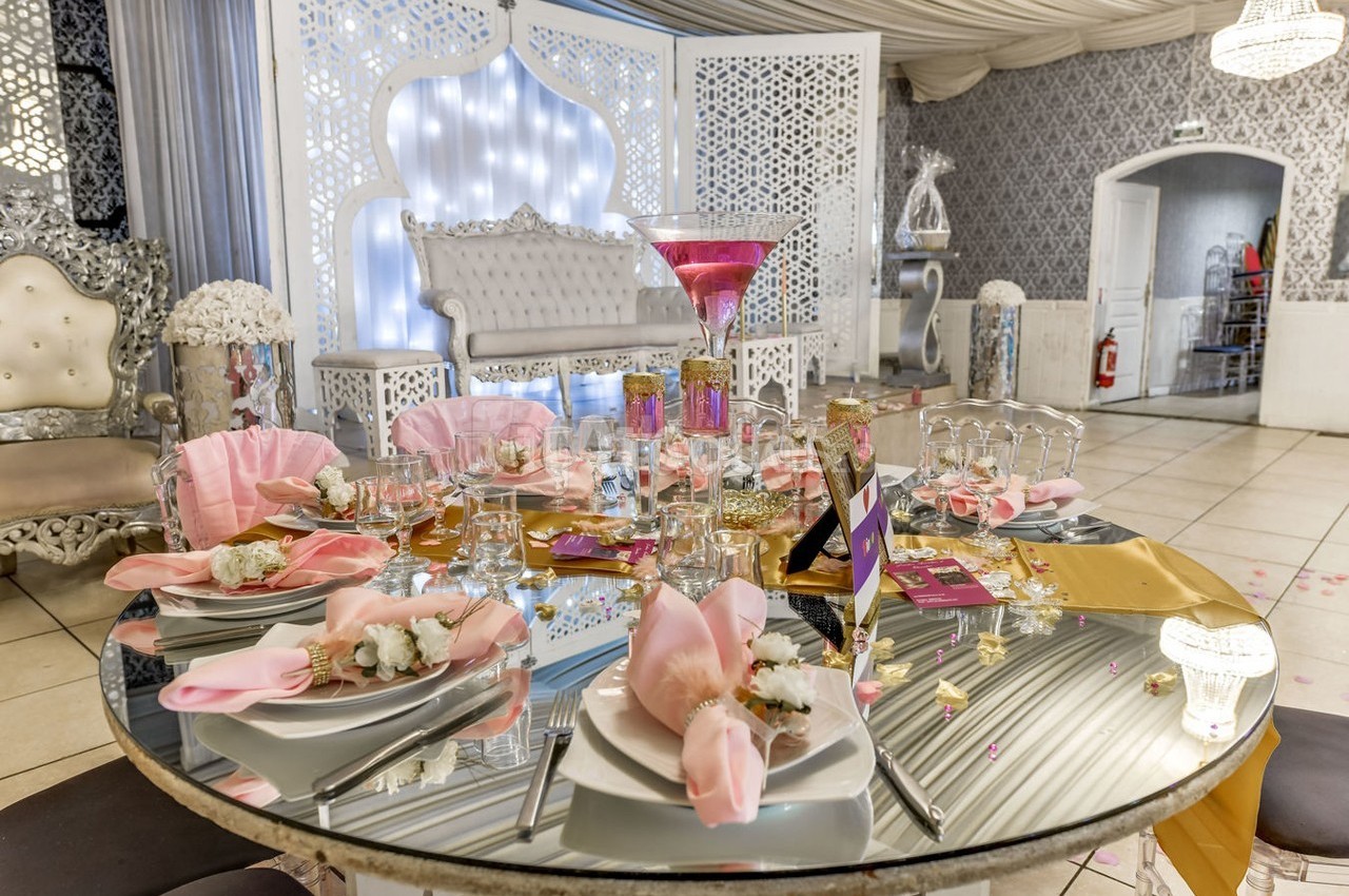 Table décorée pour un événement avec nappes roses, verres, fleurs et centre de table élégant dans une salle ornée.