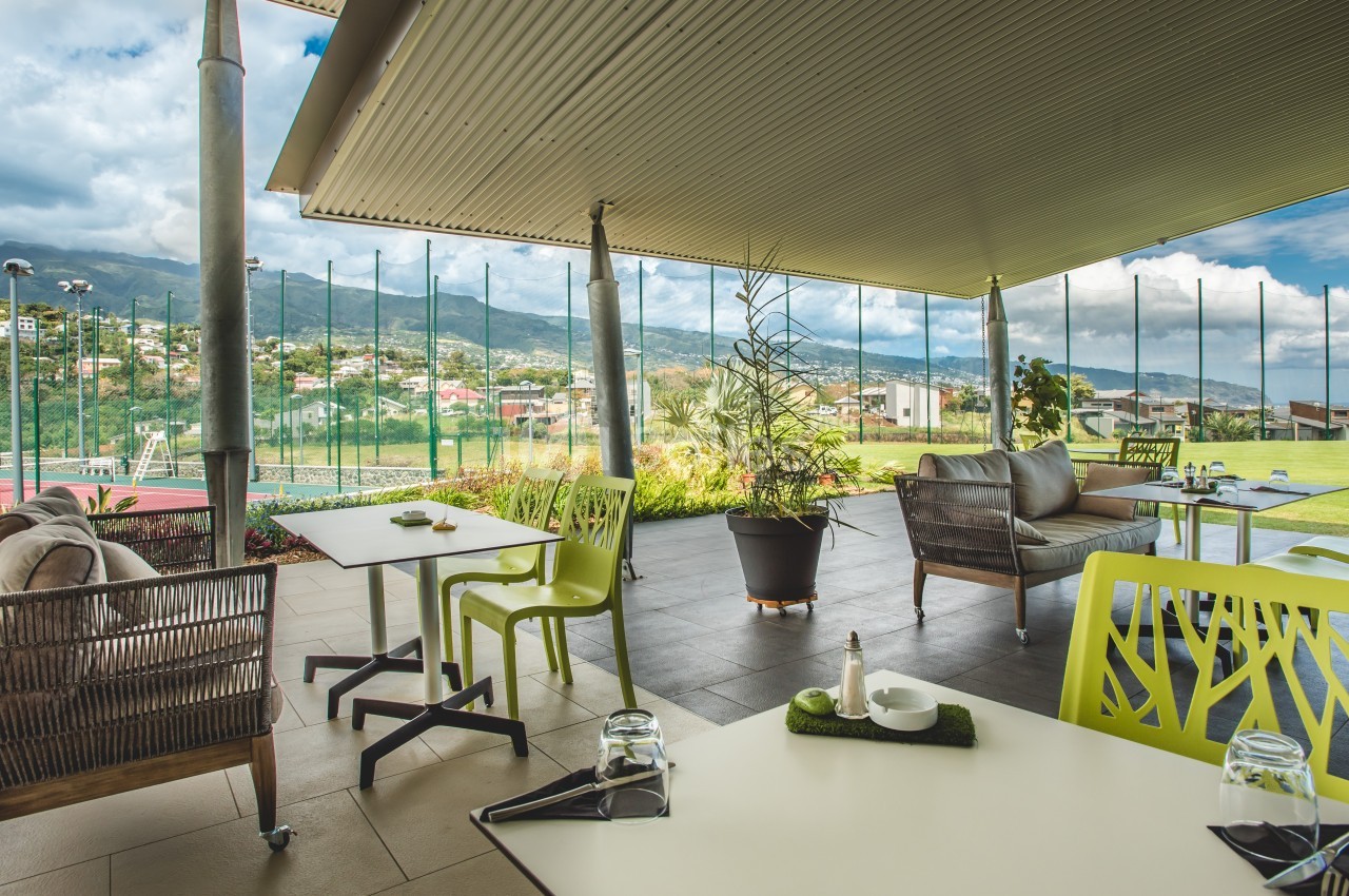 Terrasse couverte avec mobilier moderne, vue sur des collines verdoyantes et un terrain de sport en arrière-plan.