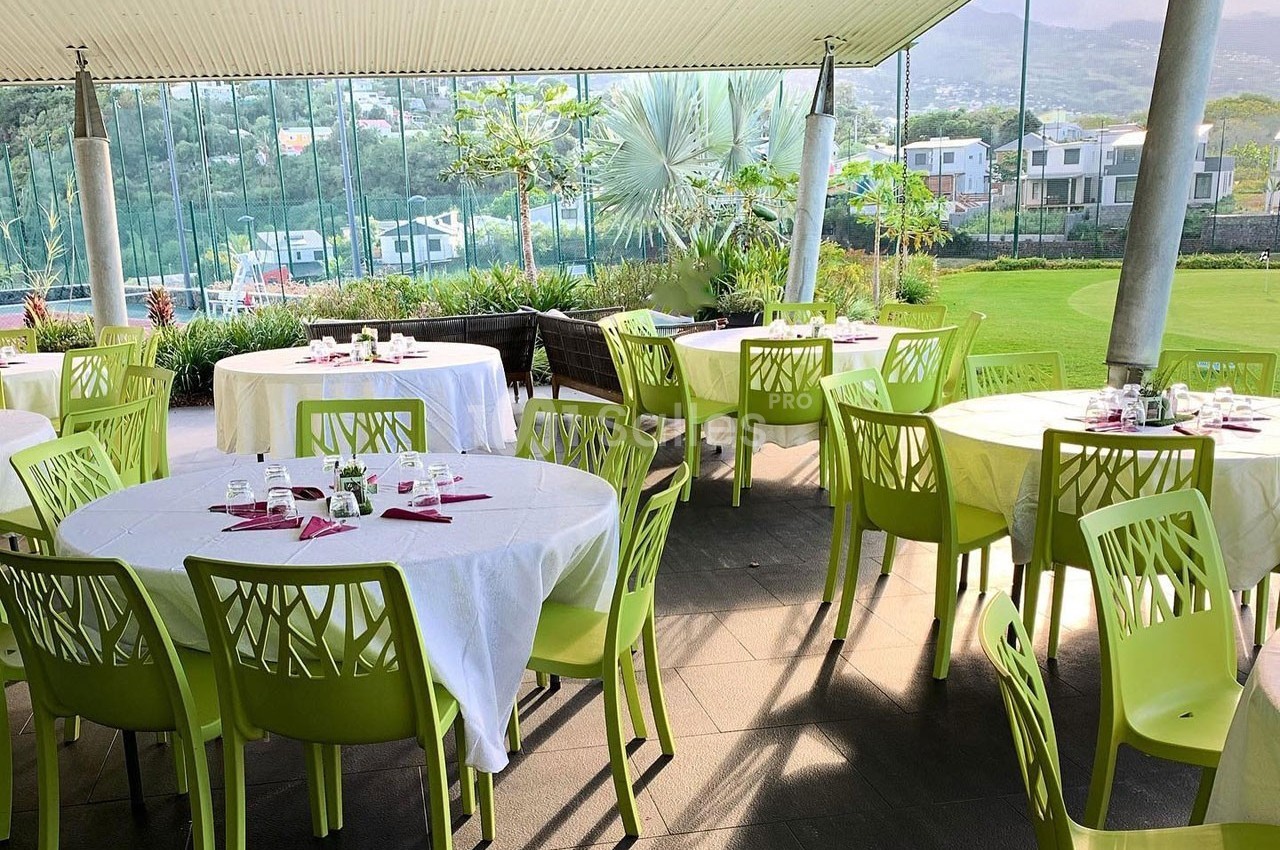 Salle à manger extérieure avec tables rondes dressées, chaises vertes et vue sur un jardin et des collines.