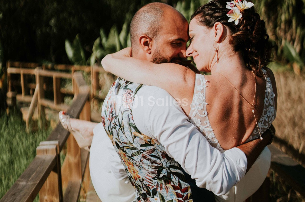 Un homme porte une femme en robe de mariée dans ses bras sur un chemin en bois entouré de végétation.