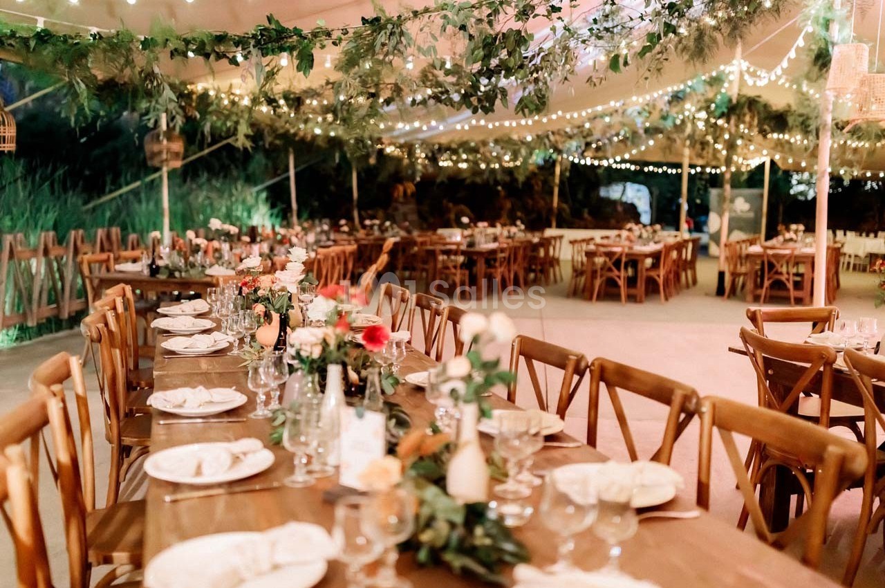 Tables en bois décorées de fleurs et de vaisselle, sous une tente éclairée par des guirlandes lumineuses.
