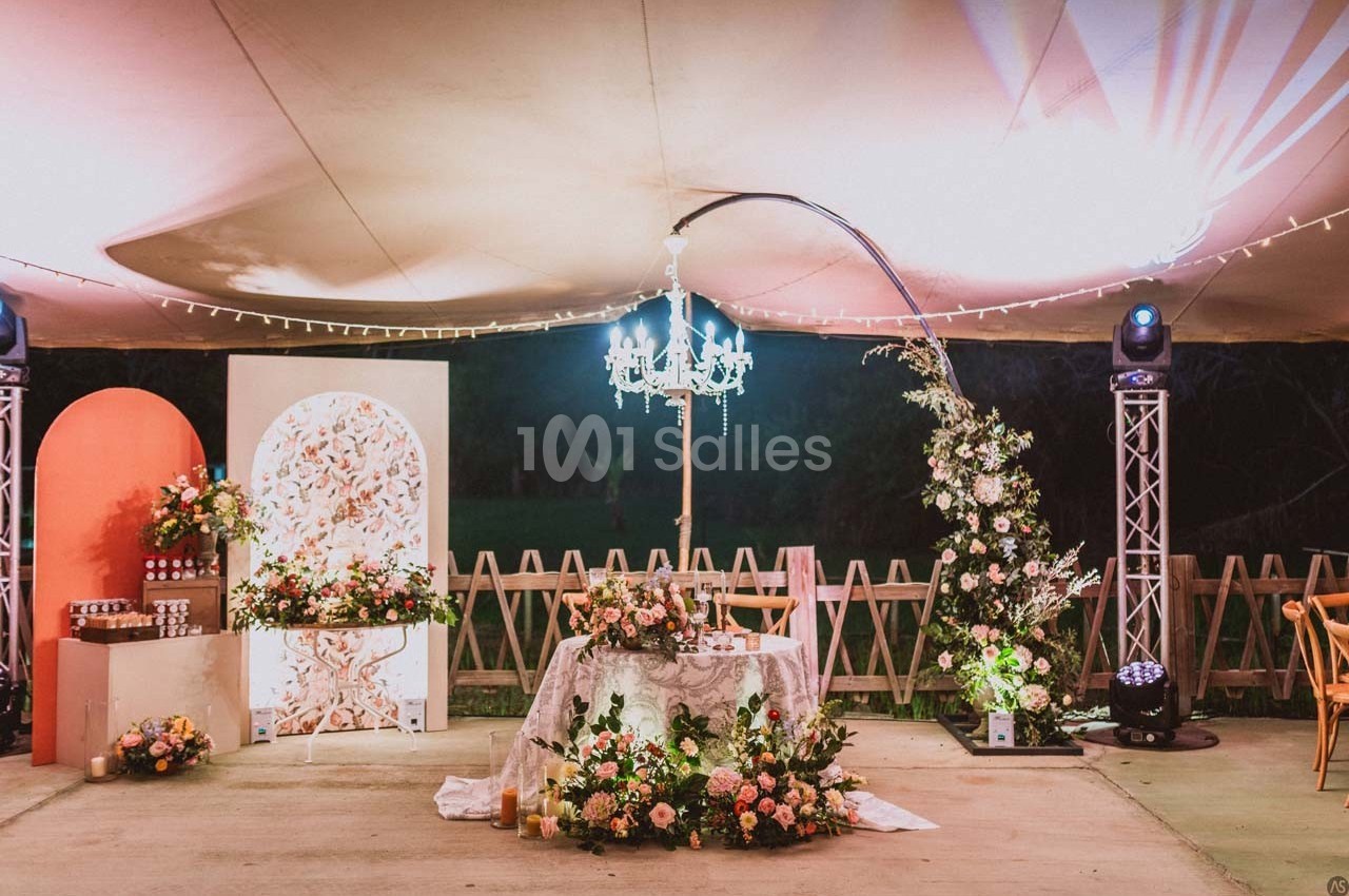 Décoration de mariage en extérieur avec table ornée de fleurs, guirlandes lumineuses et chandelier sous une tente éclairée.