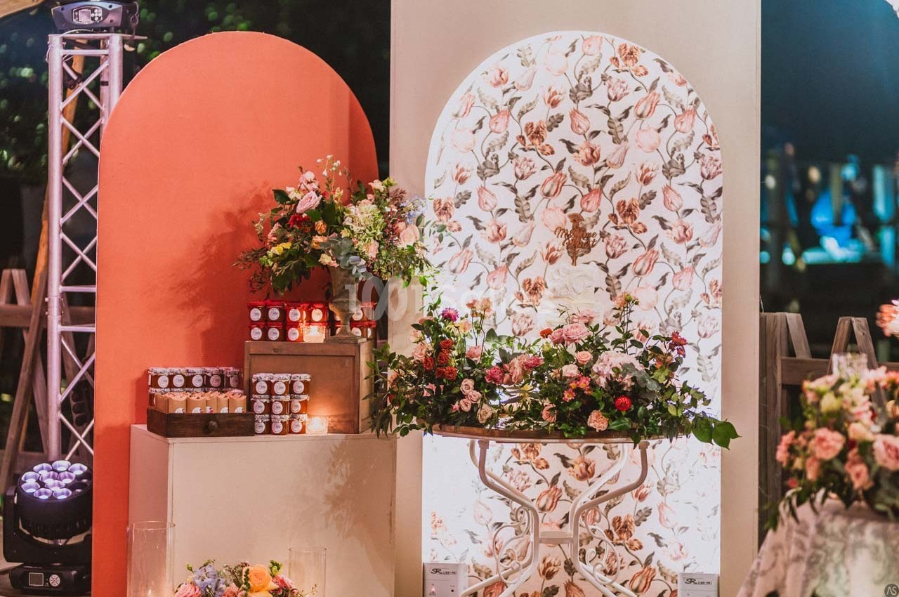 Décor floral avec table ornée de fleurs, pots de confiture alignés et panneaux colorés en arrière-plan.