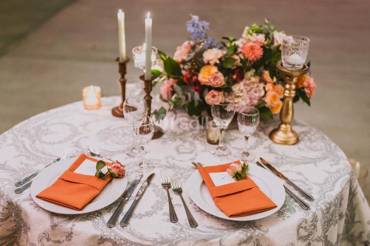 Table élégamment dressée avec nappe blanche, serviettes orange, bougies allumées et bouquet de fleurs colorées.