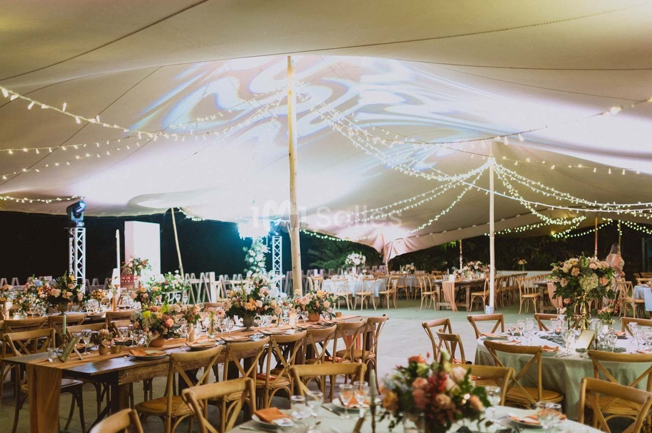 Salle de réception en plein air sous une tente illuminée, avec tables décorées de fleurs et guirlandes lumineuses.