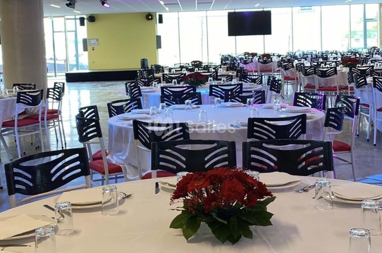 Salle de réception avec tables rondes dressées, nappes blanches, chaises noires et centres de table fleuris rouges.