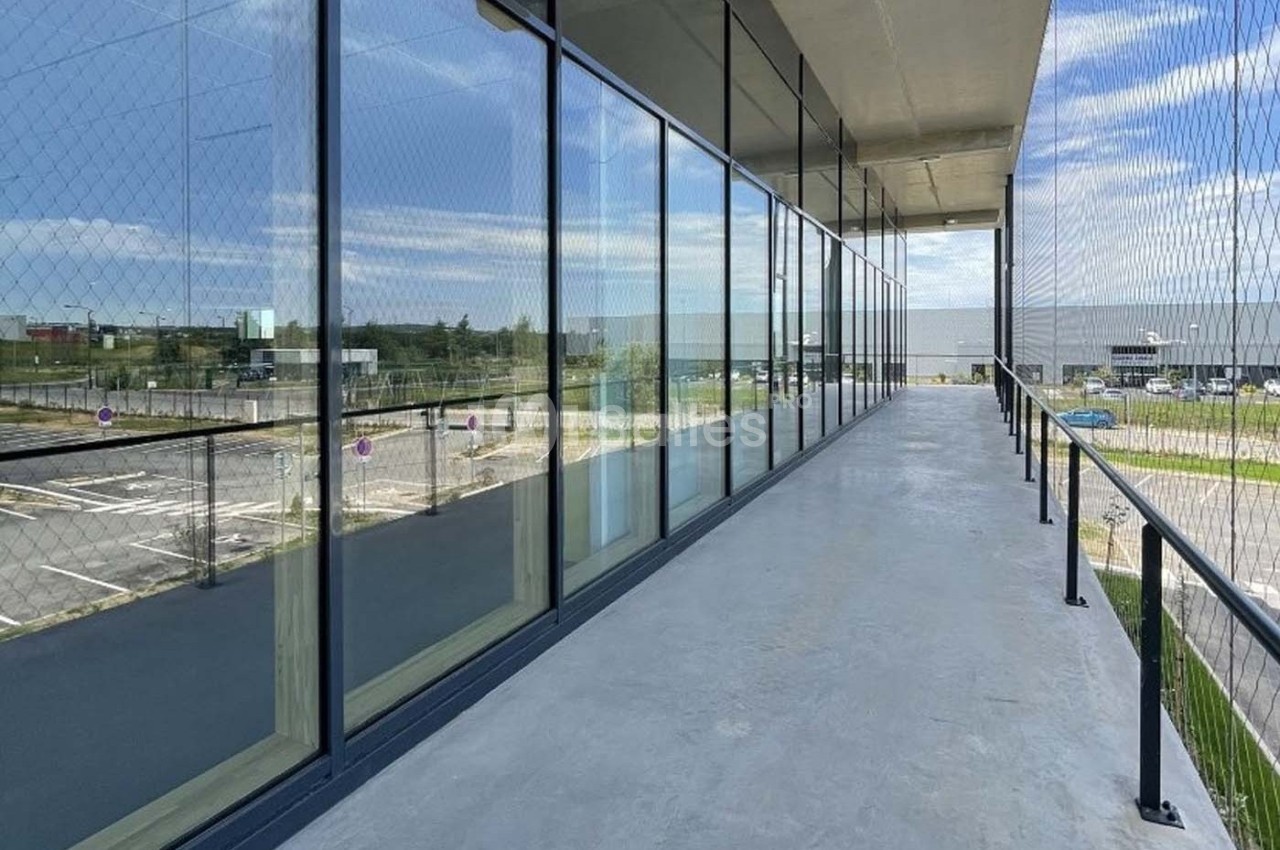 Passerelle extérieure vitrée avec vue sur un parking et un bâtiment industriel sous un ciel partiellement nuageux.