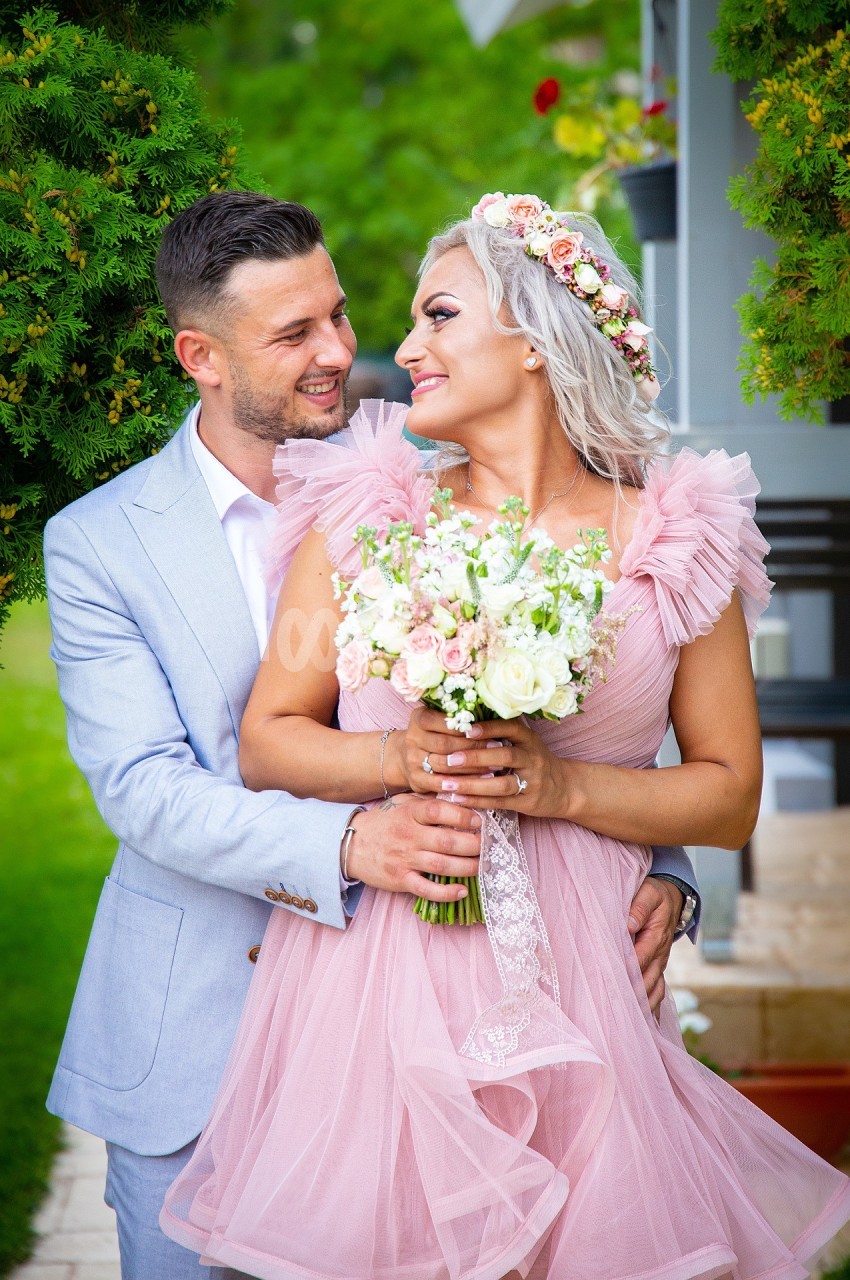 Un couple souriant en tenue élégante, la femme tenant un bouquet de fleurs, dans un jardin verdoyant.