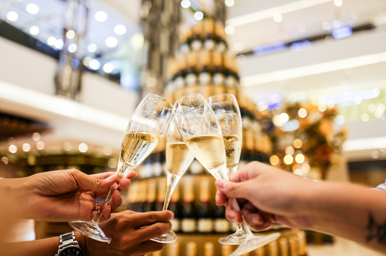 Quatre personnes trinquent avec des coupes de champagne devant une pyramide de bouteilles dans un lieu lumineux.