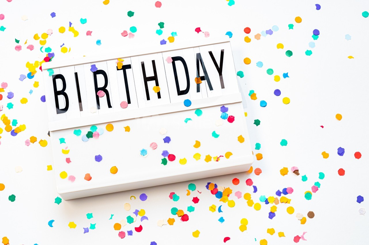Lettres formant le mot ’BIRTHDAY’ sur un panneau lumineux entouré de confettis colorés sur fond blanc.