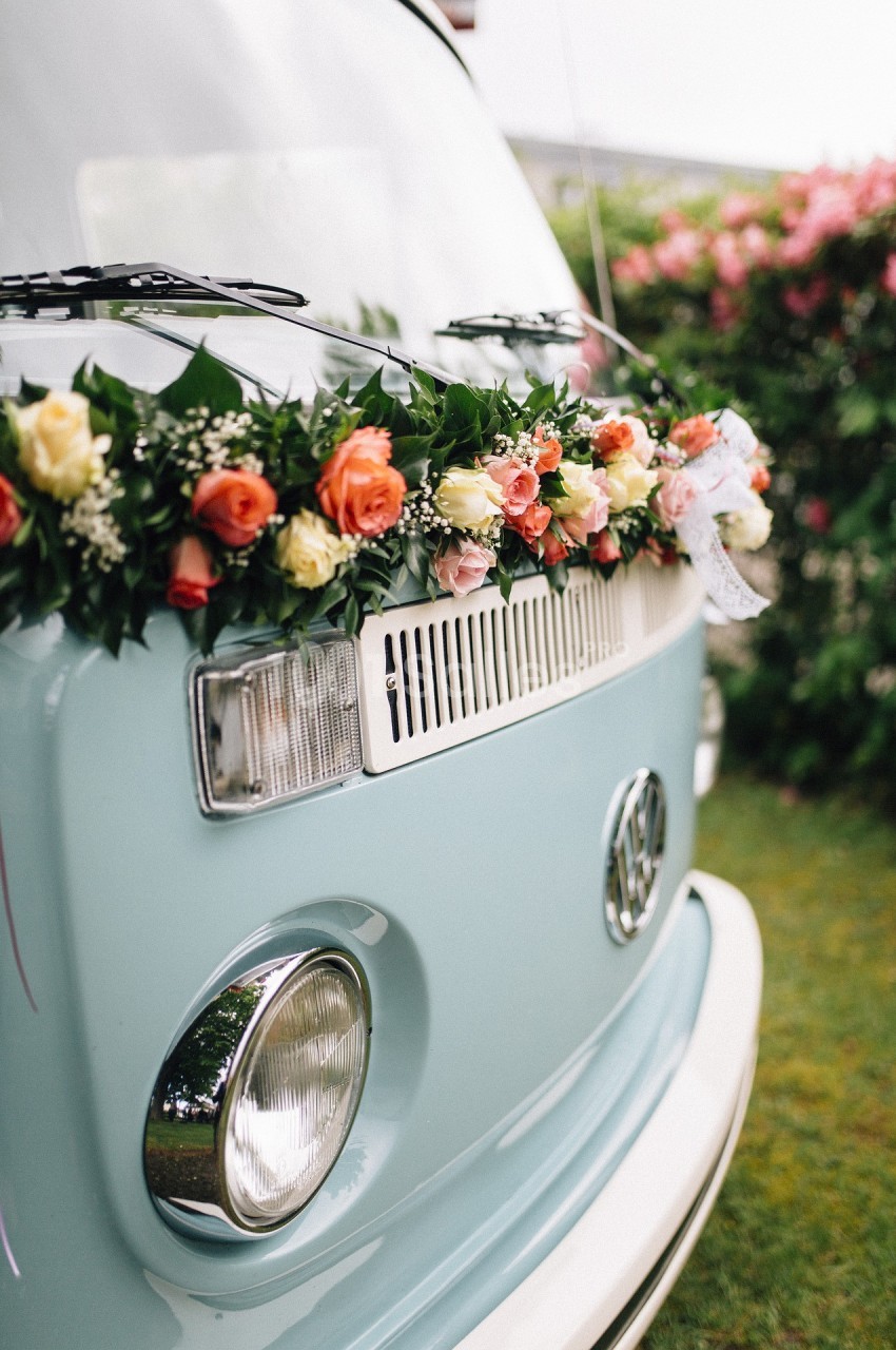 Van vintage bleu décoré d'une guirlande de fleurs colorées, garé près d'arbustes fleuris.