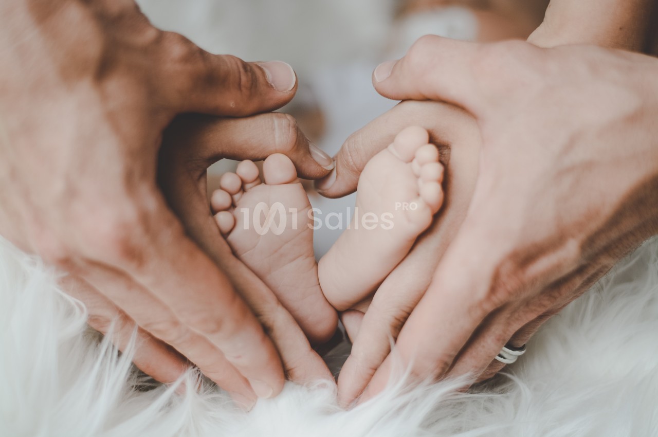 Deux mains d'adultes entourent les pieds d'un bébé, formant un cœur sur un fond de tissu blanc.