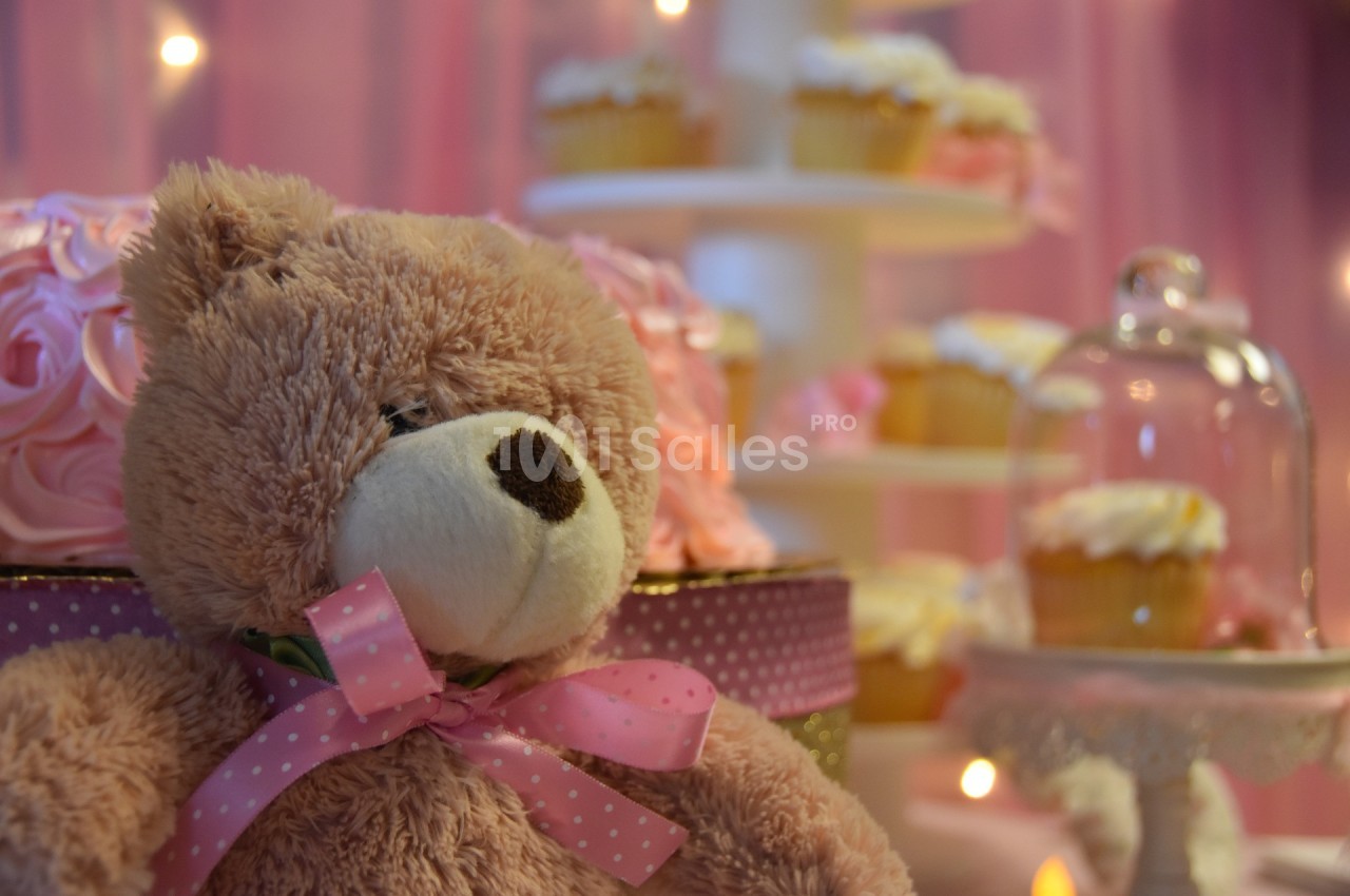 Ours en peluche avec un nœud rose devant un présentoir de cupcakes décorés dans un décor rose et lumineux.