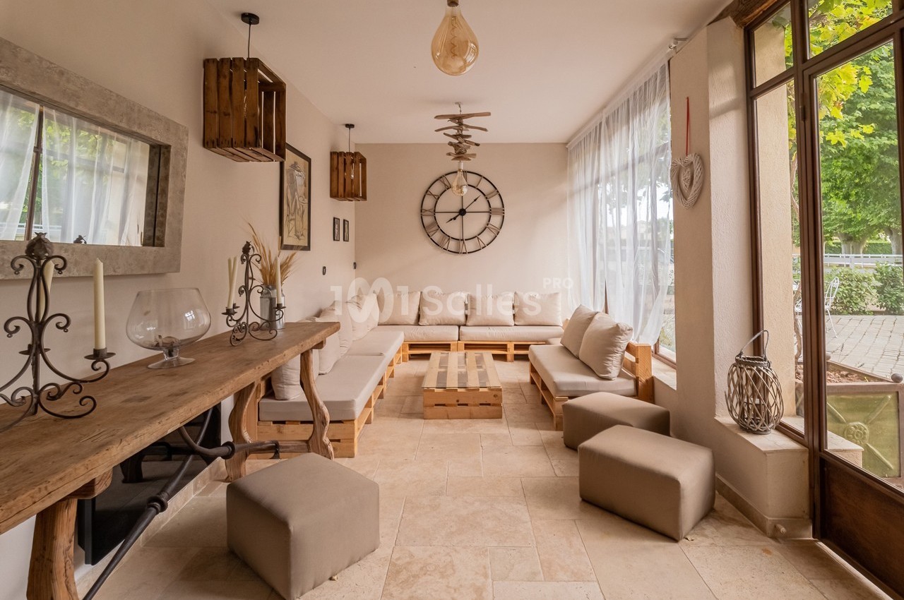 Salon lumineux avec canapé d'angle beige, table basse en bois, décoration murale et grandes fenêtres donnant sur l'extérieur.