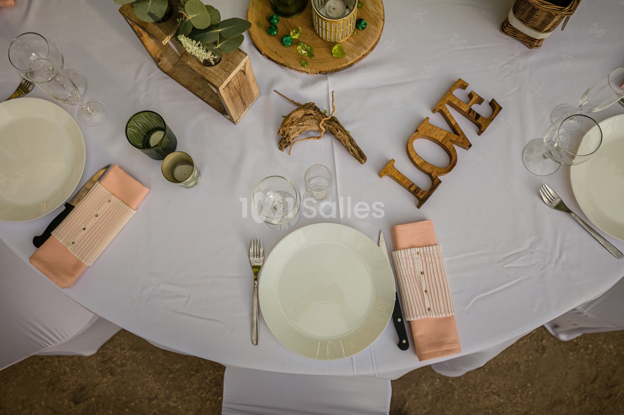 Table dressée avec nappes blanches, assiettes, couverts, serviettes roses et décorations en bois sur le thème de l'amour.