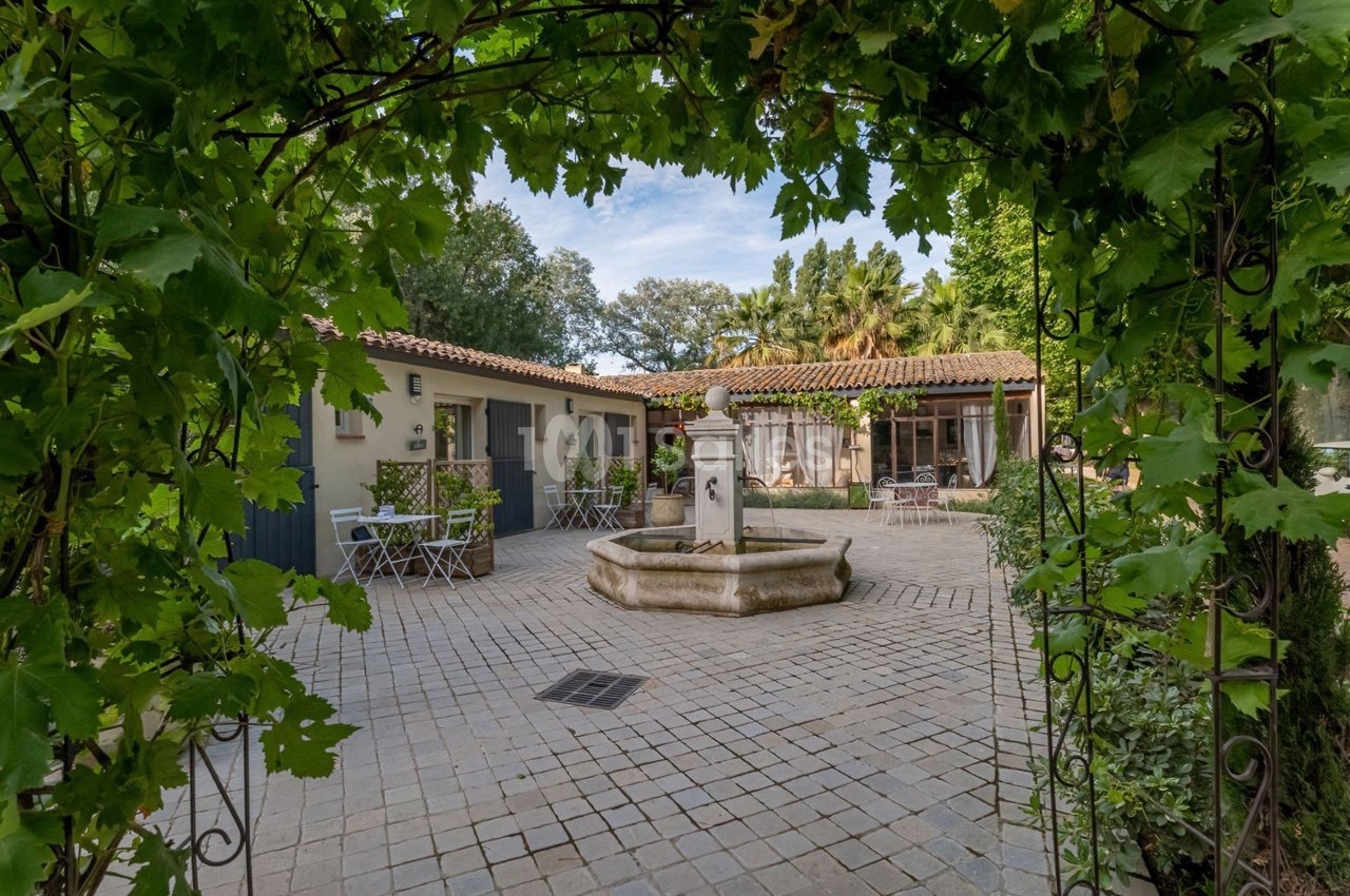 Cour pavée avec une fontaine centrale, entourée de bâtiments à toit en tuiles et de végétation verdoyante.