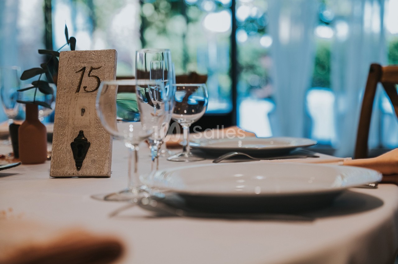 Table dressée avec des assiettes, verres et serviettes, décorée d'un numéro de table en bois marqué ’15’.
