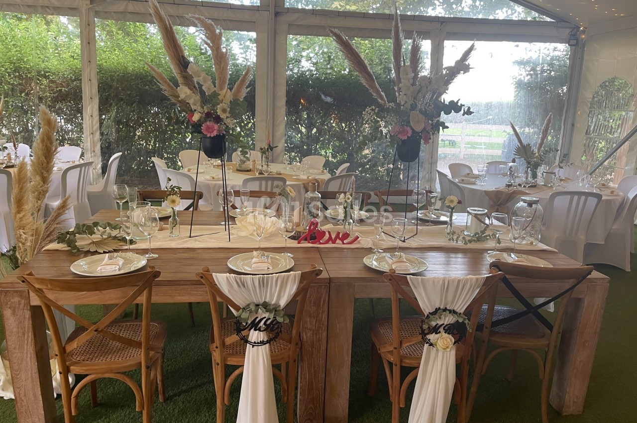 Table de mariage décorée avec des fleurs, des nappes blanches et des chaises ornées de voilages dans une salle lumineuse.