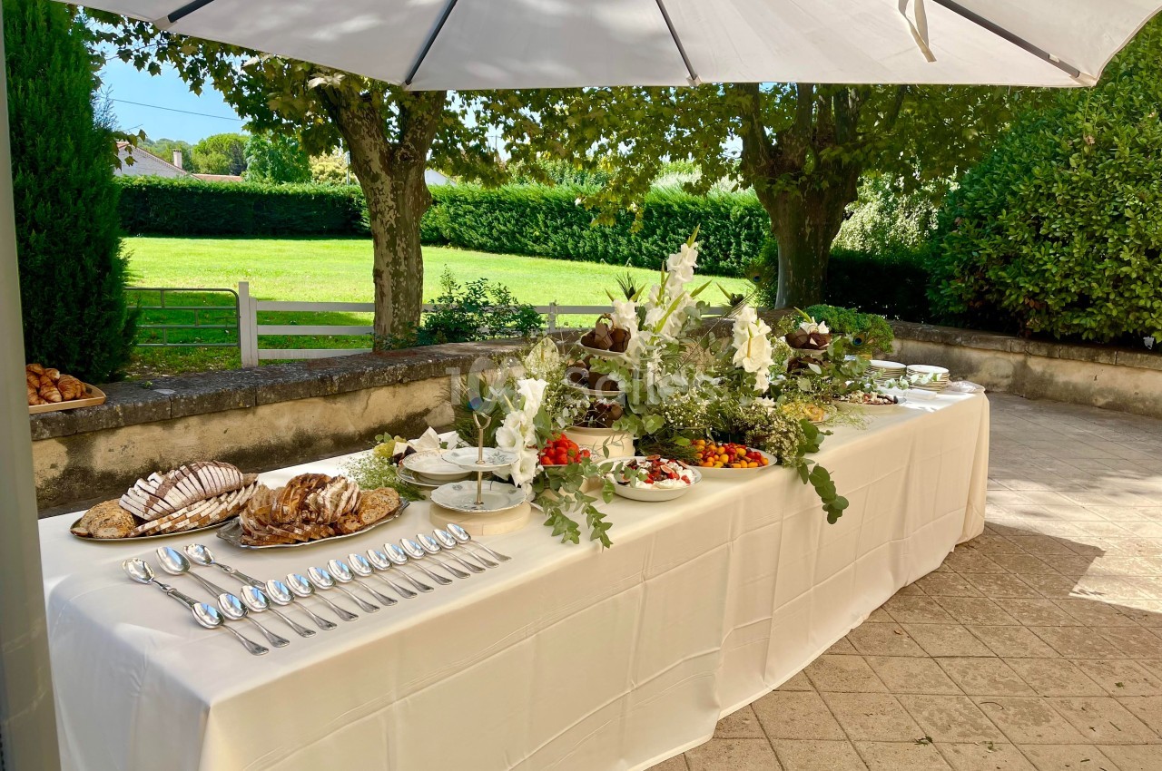 Table dressée en extérieur avec plats variés, décorée de fleurs blanches, sous un grand parasol près d'un jardin.