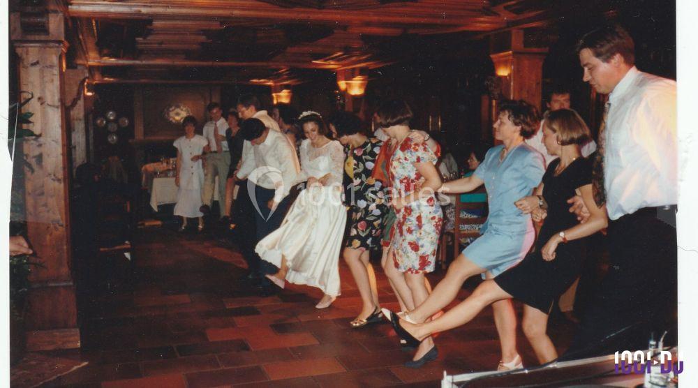 Des personnes, dont une mariée en robe blanche, dansent en ligne dans une salle au décor en bois.