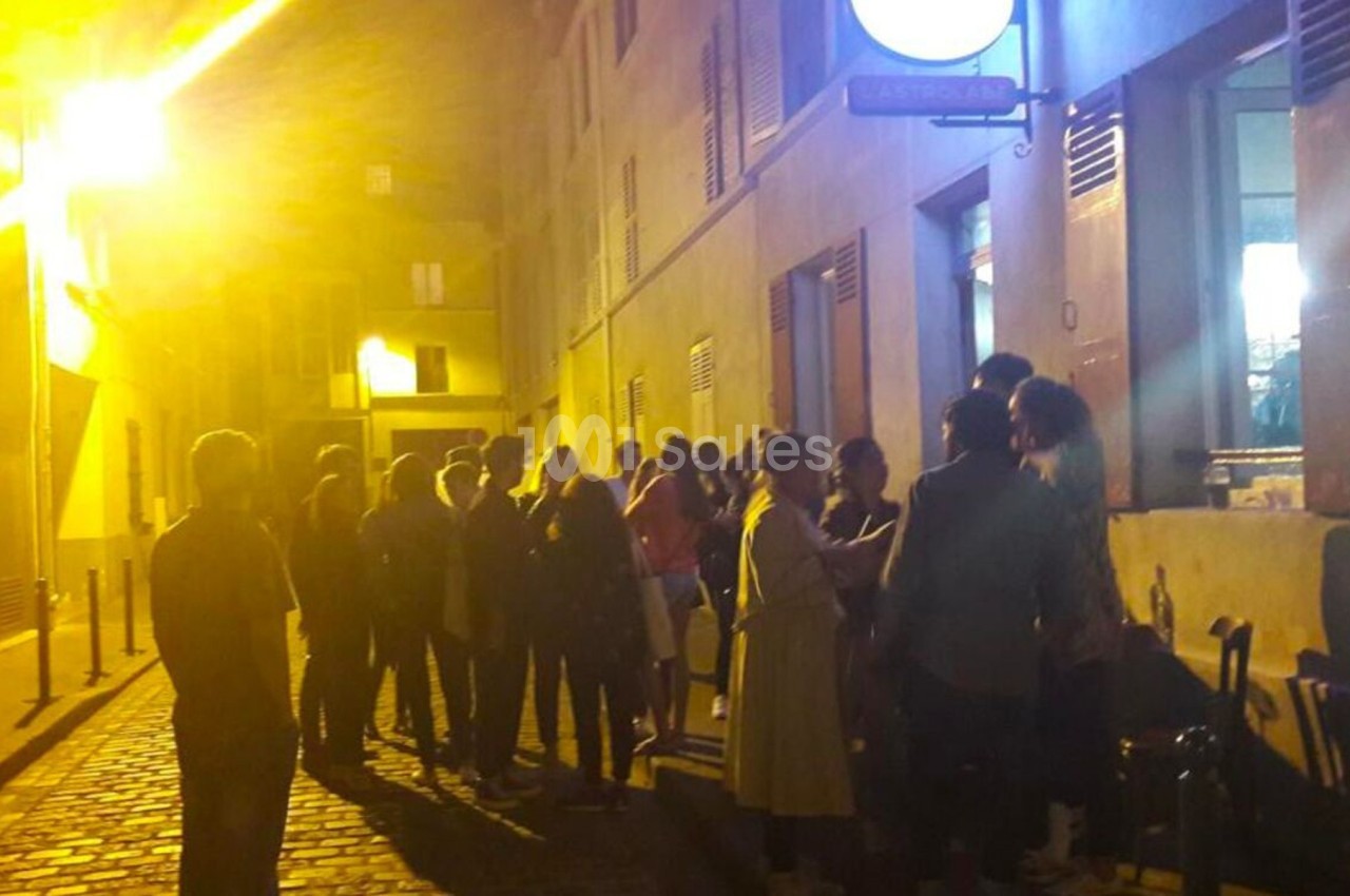 Groupe de personnes faisant la queue devant un bâtiment éclairé, dans une rue pavée en soirée.