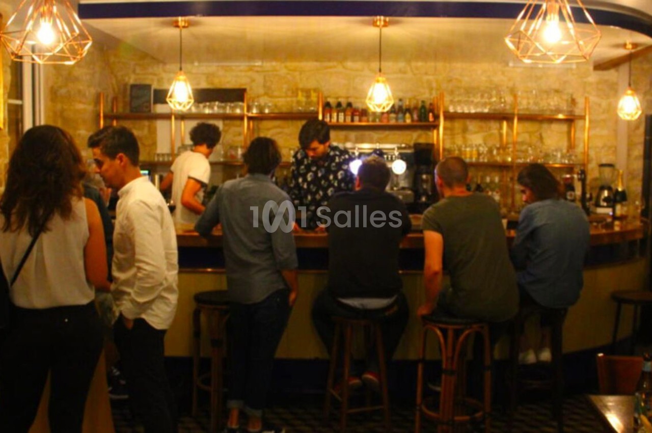 Des personnes discutent et commandent au comptoir d'un bar éclairé par des suspensions lumineuses.