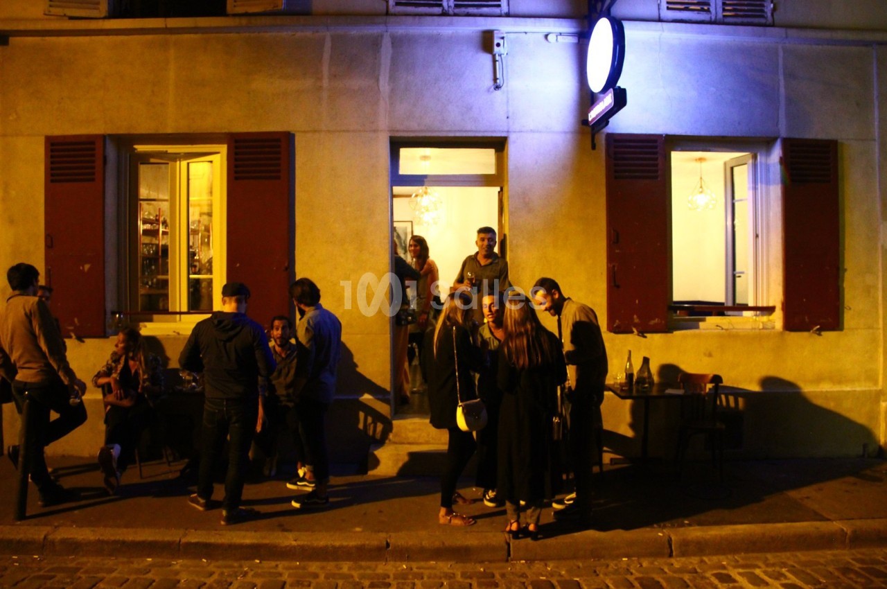 Groupe de personnes discutant devant un café éclairé en soirée, avec des fenêtres ouvertes et une lumière bleutée.