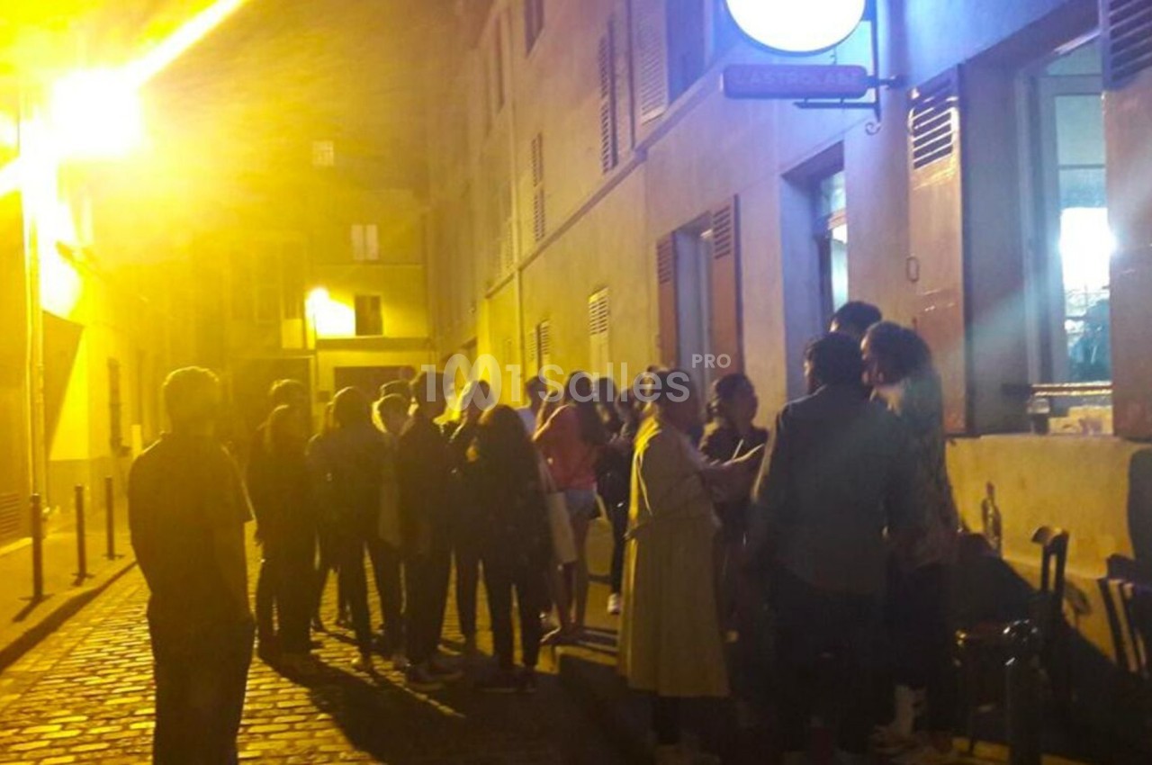 Groupe de personnes faisant la queue devant un bâtiment éclairé, dans une rue pavée en soirée.