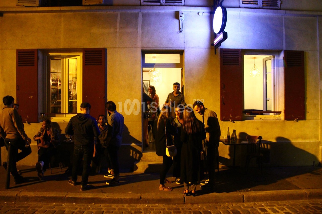 Groupe de personnes discutant devant un café éclairé en soirée, avec des fenêtres ouvertes et une lumière bleutée.