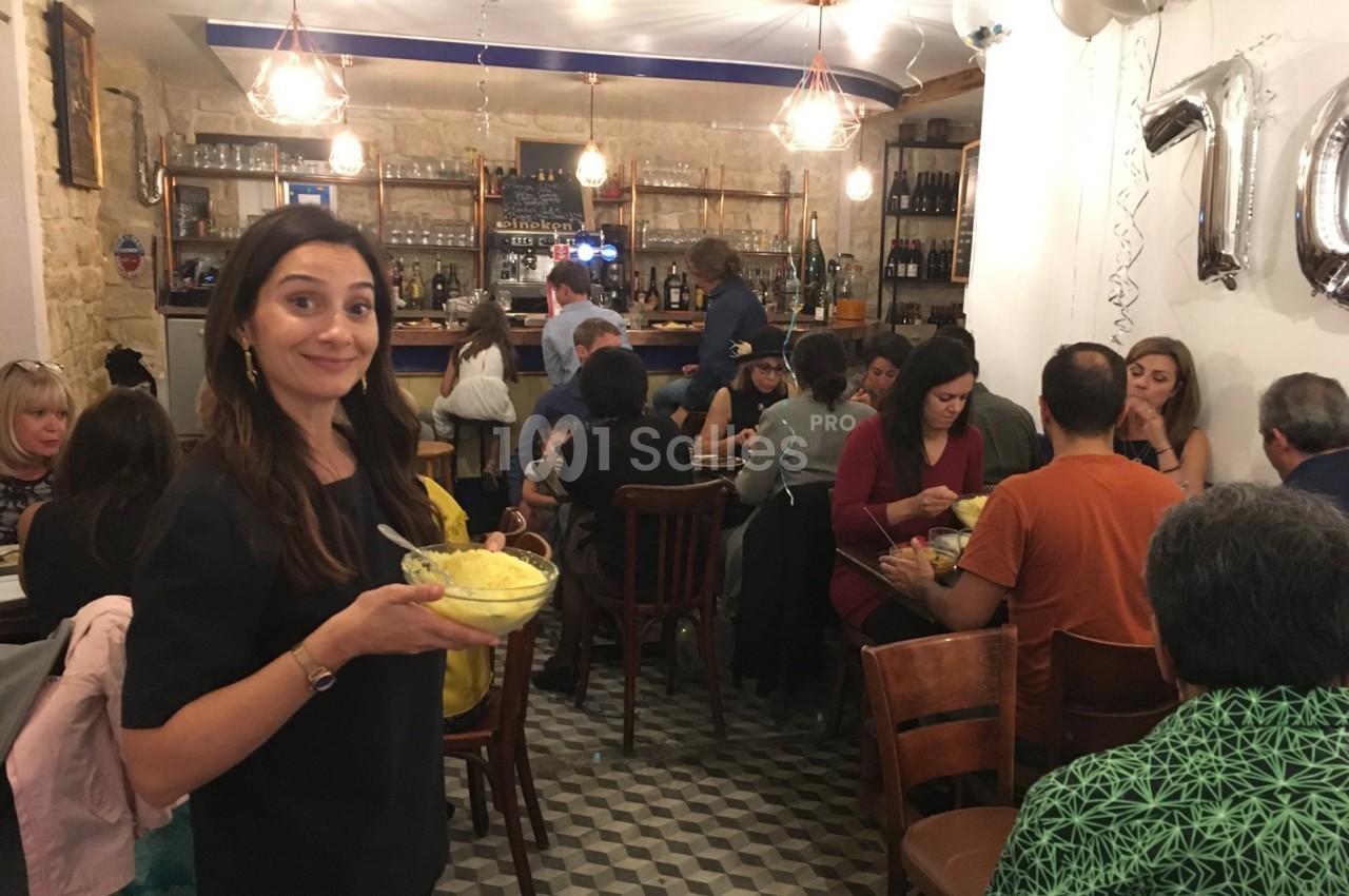 Une femme souriante tient un plat dans un restaurant animé avec des convives assis à des tables.
