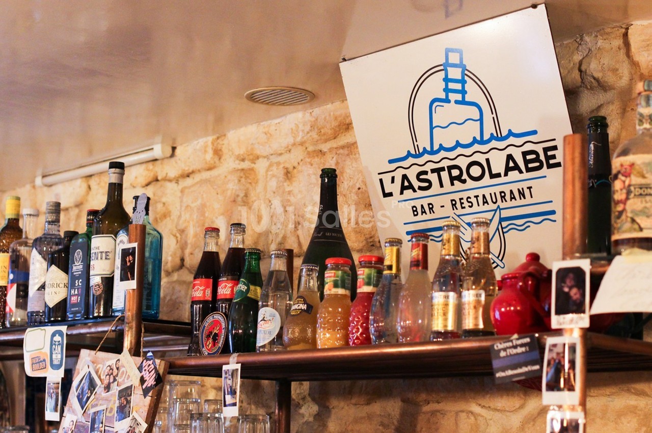 Bouteilles variées alignées sur une étagère en bois dans un bar avec une enseigne ’L'Astrolabe’ en arrière-plan.