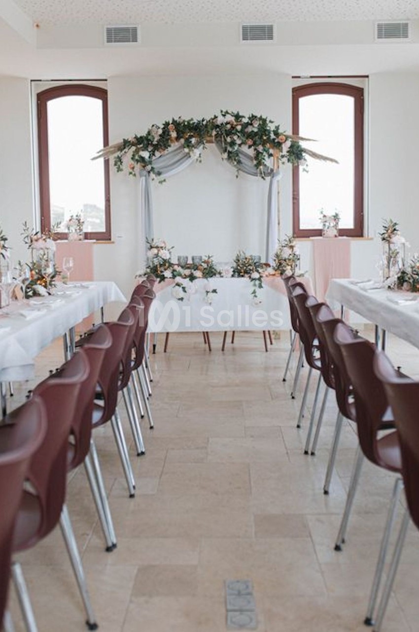 Salle décorée pour un mariage avec une arche florale, tables alignées et chaises bordeaux.