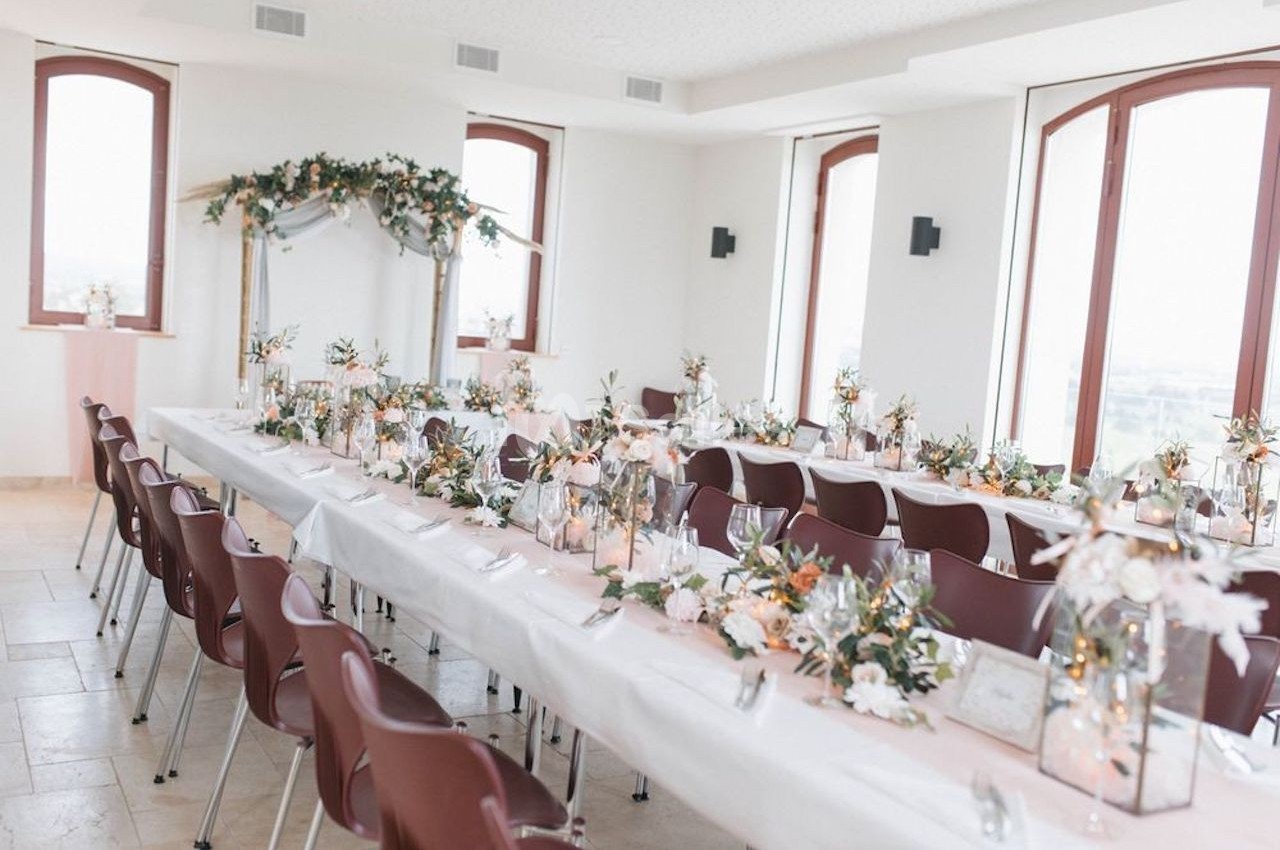 Salle lumineuse décorée pour un mariage, avec longues tables ornées de fleurs et chaises alignées.