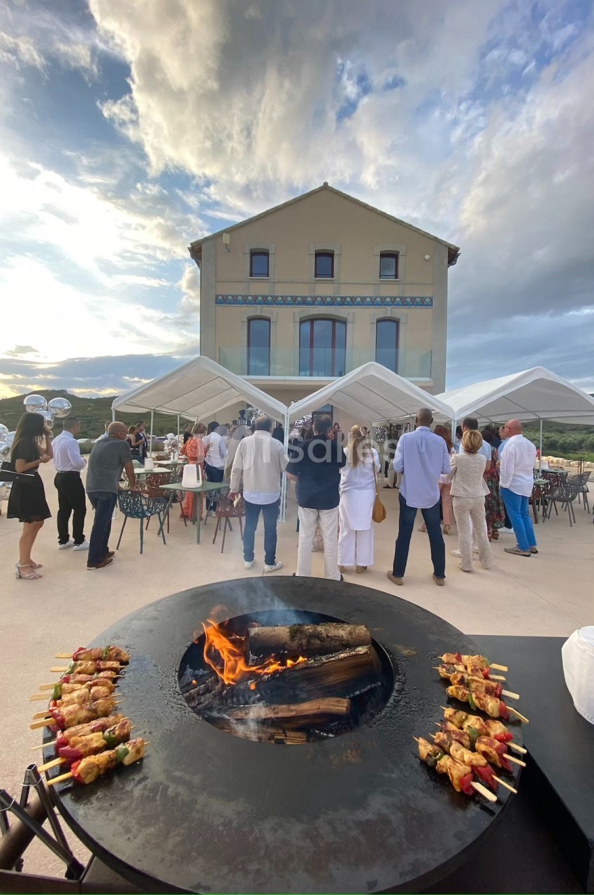 Personnes rassemblées devant une grande maison avec des tentes blanches, un brasero avec brochettes au premier plan.