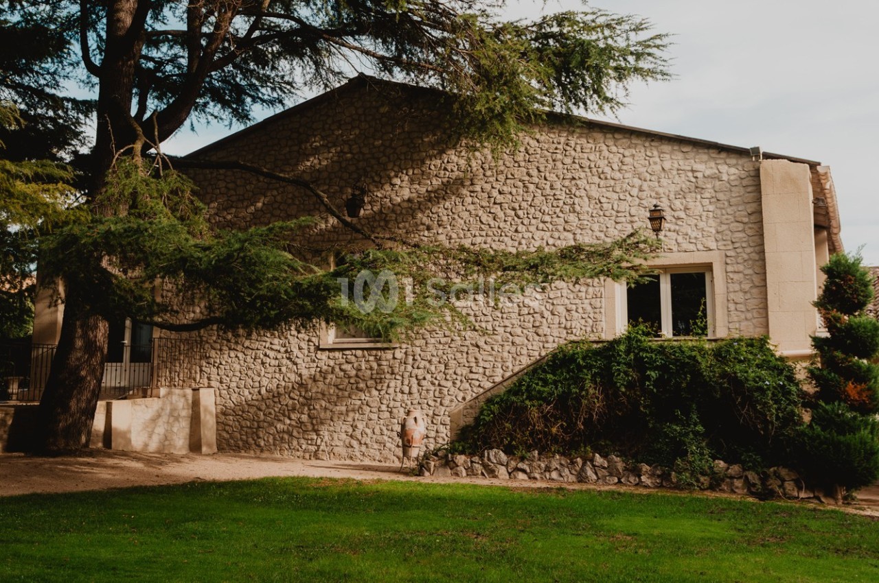 Bâtiment en pierre entouré d'arbres et de verdure, avec une façade simple et des fenêtres rectangulaires.