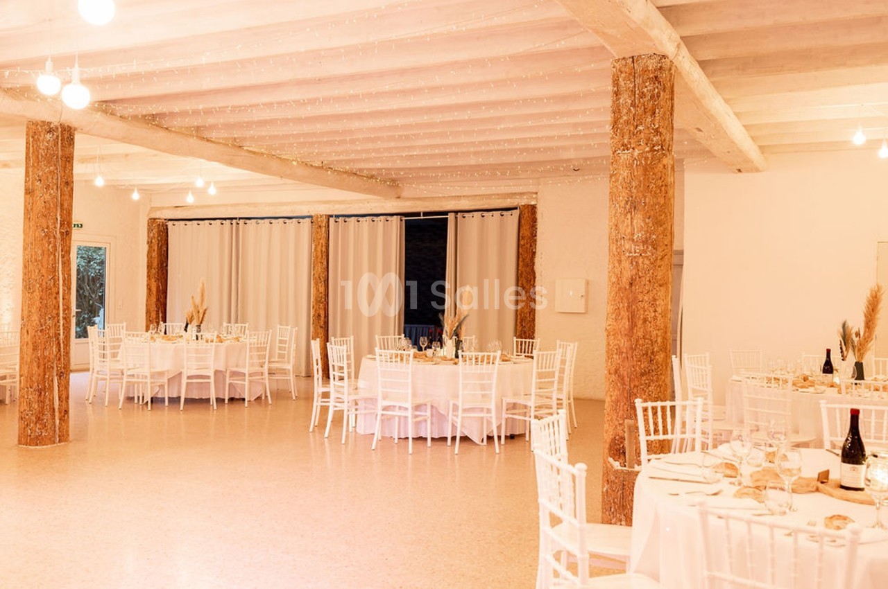 Salle de réception lumineuse avec tables rondes dressées, chaises blanches et colonnes en bois brut.